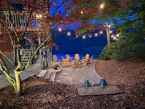 An outdoor seating area with Adirondack chairs and a fire pit, surrounded by trees and string lights.