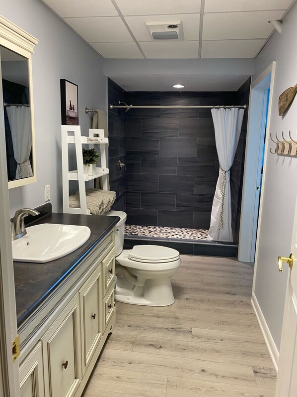 A bathroom with a white sink, a toilet, a shower with a dark tiled wall, and a light-colored wooden floor.