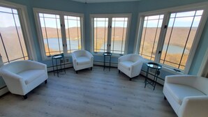 Une pièce comprenant quatre fauteuils blancs disposés autour d’une table basse centrale, de grandes fenêtres donnant sur un paysage désertique et un ventilateur de plafond.