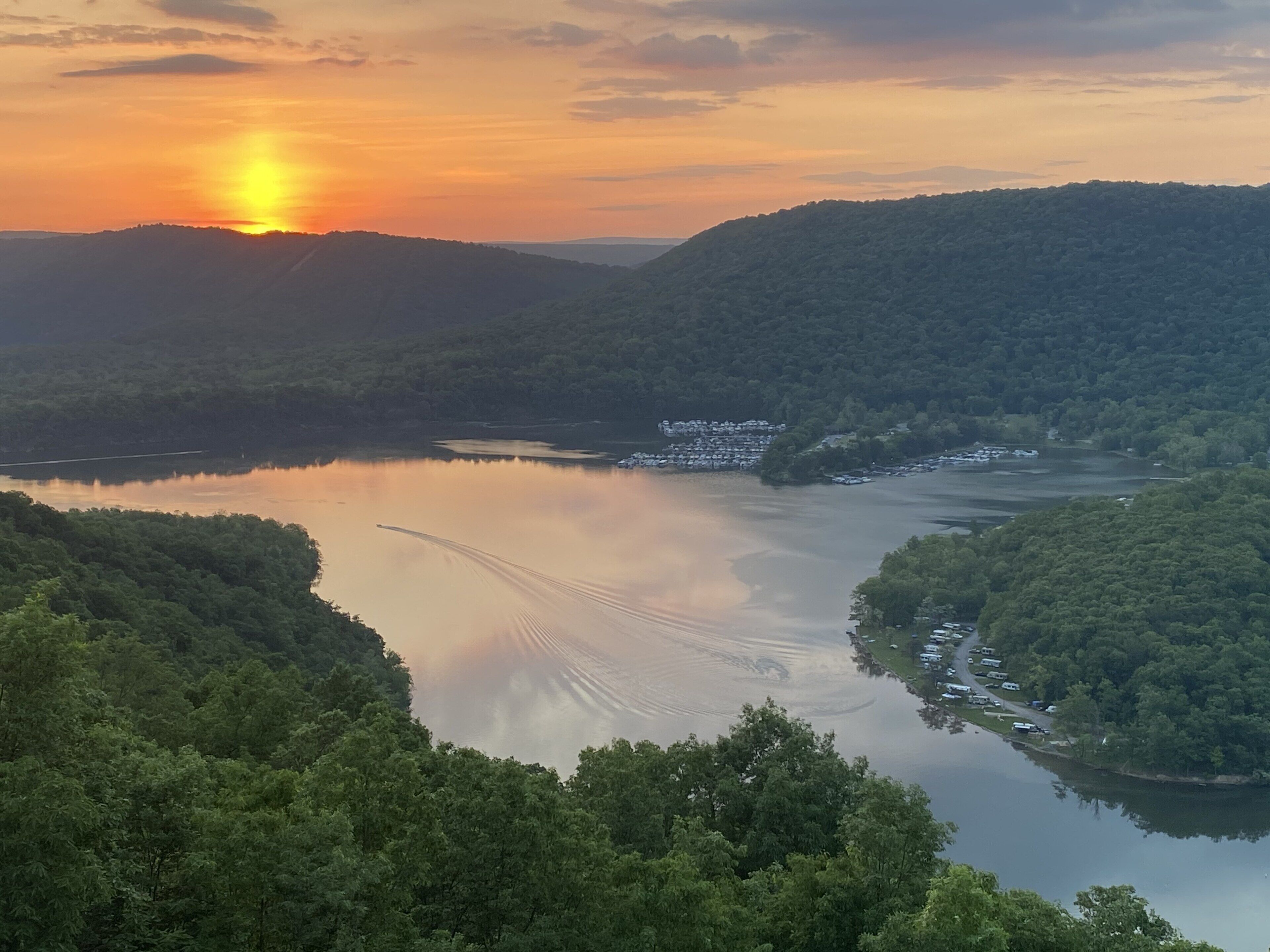 Ein See, umgeben von bewaldeten Hügeln, mit freiem Blick auf den Sonnenuntergang.