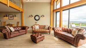 A spacious living room with three leather sofas and a matching ottoman, wooden flooring, large windows with a view, and a chandelier. - Valley View Lodge Sleeps 8 With Hot Tub (Preston)