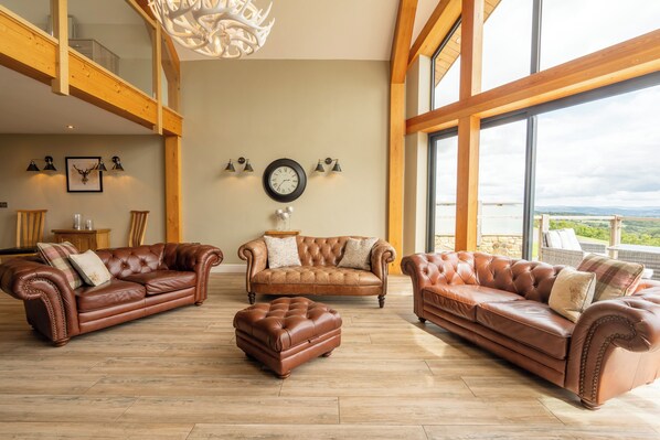 A spacious living room with three leather sofas and a matching ottoman, wooden flooring, large windows with a view, and a chandelier. - Valley View Lodge Sleeps 8 With Hot Tub (Preston)