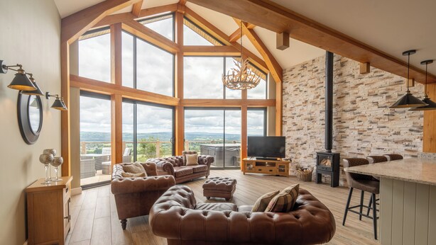 A spacious living room with a large brown leather sofa, a wooden coffee table, and a stone wall. A large window offers a view of the outdoors. - Valley View Lodge Sleeps 8 With Hot Tub (Preston)