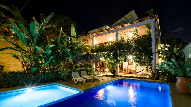 A swimming pool with lounge chairs and an umbrella, surrounded by lush greenery and a building with outdoor lighting. - Los Gallos - Second Level Apartment (Lo de Marcos)