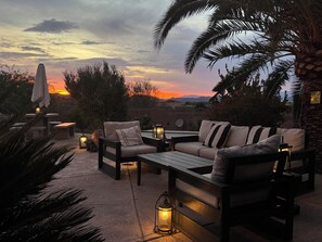 An outdoor seating area with a sofa and chairs, surrounded by palm trees and a sunset view. - Legendary Desert Oasis -Heated Pool + Spa -Golf -Luxury 5BR Retreat Sleeps 12  (Phoenix)