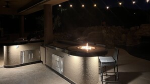 Uma área de bar ao ar livre com churrasqueira integrada, assentos e um fundo de pedra.