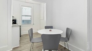 Une petite pièce moderne avec une table ronde et quatre chaises, une fenêtre avec des stores et un coin cuisine.
