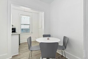 Quarto pequeno e moderno com uma mesa redonda e quatro cadeiras, uma janela com persianas e um espaço com cozinha compacta.