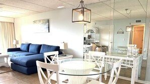 A living room with a blue sofa, a glass dining table with white chairs, and a chandelier. - Amazing Pool/Oceanfront Views from this Second Floor Condo at Amelia by the Sea! (Fernandina Beach)