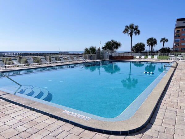 Una piscina rectangular con una marca de profundidad de 1 metro 17, rodeada de sillas de descanso y palmeras, con vista al océano y un edificio al fondo.