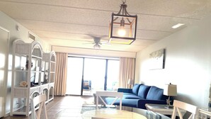 A spacious living area with a blue sofa, a round glass table, and white chairs. A ceiling fan and a hanging light fixture are visible. - Amazing Pool/Oceanfront Views from this Second Floor Condo at Amelia by the Sea! (Fernandina Beach)