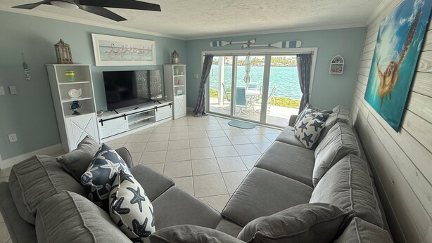A living room with a large gray sectional sofa, a flat-screen TV, and a ceiling fan. There is a sliding glass door leading to a balcony with a view of the water. - Dolphin & Manatee At Your Back Door!! (Treasure Island)