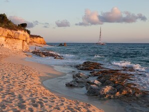 Una spiaggia con un litorale roccioso, una barca a vela in lontananza e una scogliera ricoperta di vegetazione.
