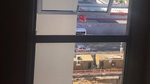 Une vue à travers la fenêtre d’un immeuble, avec un ciel dégagé, une étendue d’eau au loin et un chantier de construction en contrebas.