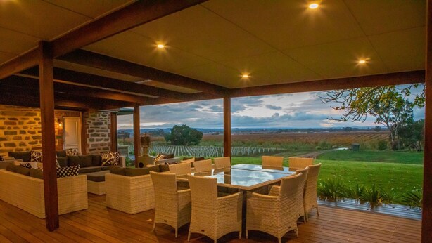 Un área de estar al aire libre con mobiliario de mimbre y una mesa con cubierta de vidrio, con vista a un viñedo.