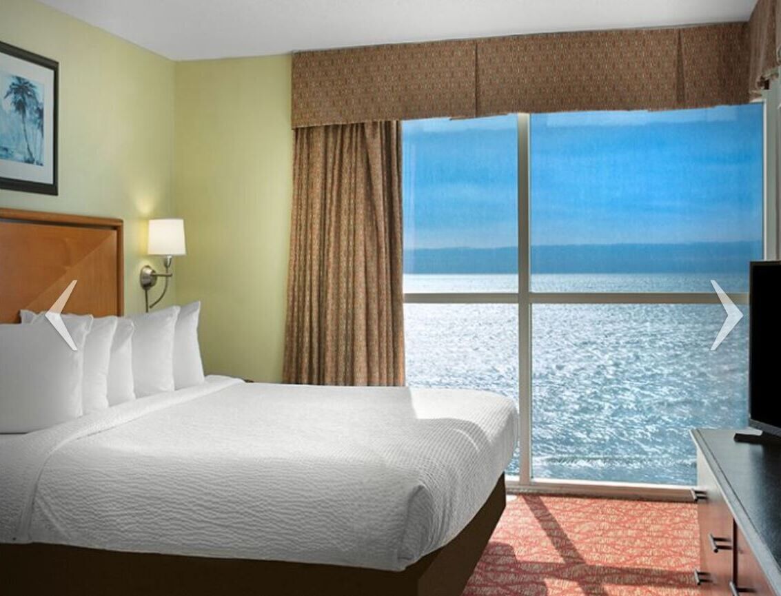 A hotel room with a large bed, a television, a window with a view of the sea, and a lamp.