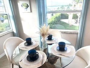 A round glass table with four blue cups and saucers, a vase with dried plants, and a view of a garden through a window.
