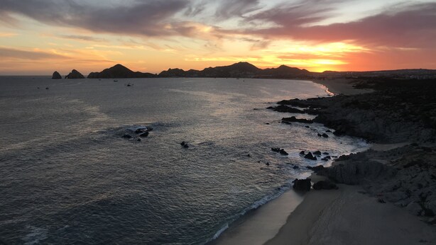 Paisaje costero con playa, costa rocosa y montañas distantes al atardecer.