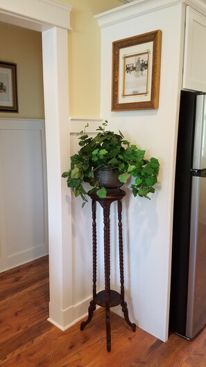 Un coin de pièce avec un plancher en bois, un réfrigérateur noir, un tableau encadré accroché au mur et une plante en pot posée sur un support en bois.
