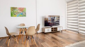 Un salon moderne avec un téléviseur, une table à manger avec des chaises, un tableau abstrait aux couleurs vives et une grande fenêtre avec des stores.