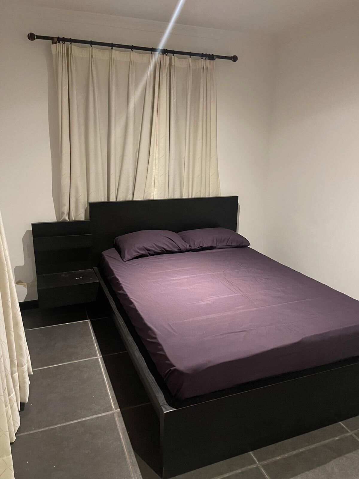 A single bed with a black frame and a purple mattress, a nightstand, and a curtain.