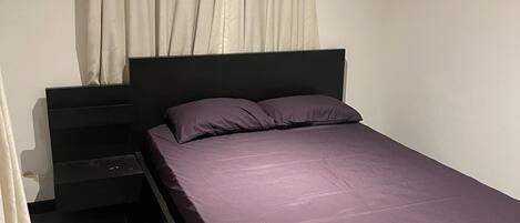 A single bed with a black frame and a purple mattress, a nightstand, and a curtain.