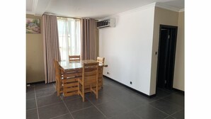 A dining area with a glass-top table and wooden chairs, a window with curtains, and a door with an air conditioning unit on the wall. - Remarkable 2-Bed Apartment in Accra (Accra)