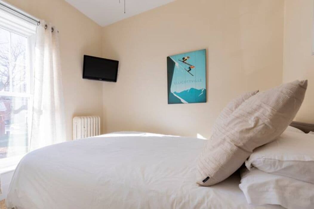 Ein Schlafzimmer mit Bett, einem Fenster mit Vorhängen, einem an der Wand montierten Fernseher und einem Poster.