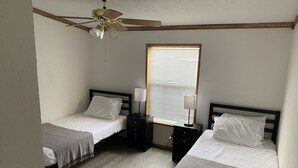 Quarto com duas camas de solteiro, ventilador de teto, janela com persianas e mesa de cabeceira com abajur.