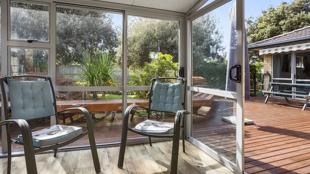 Eine überdachte Terrasse mit zwei Stühlen und einem Tisch, große Fenster mit Blick auf einen Garten und ein Haus.