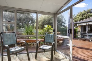 Alpendre coberto com duas cadeiras e uma mesa, grandes janelas proporcionando vista para um jardim e uma casa.