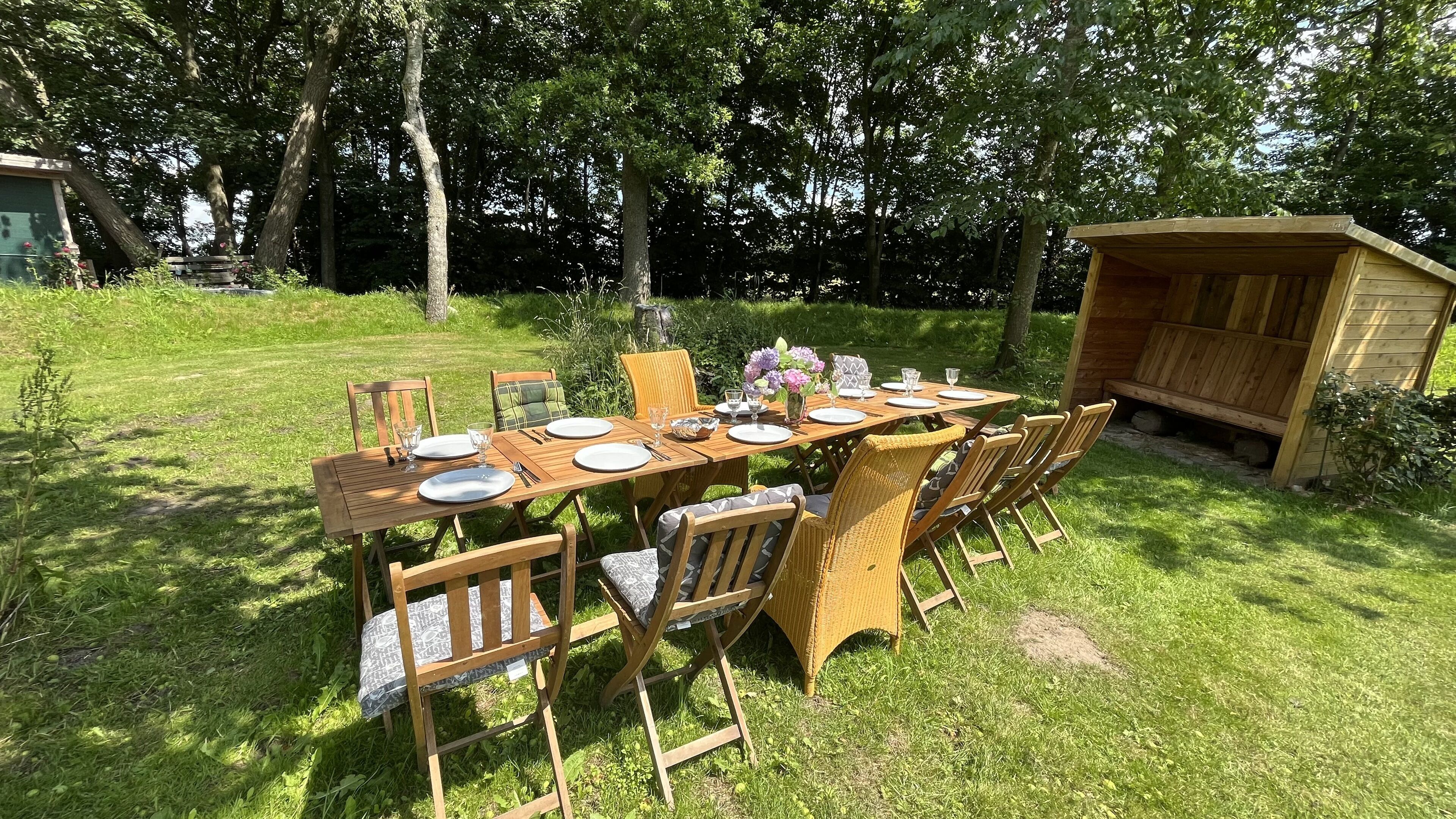 Mesa de madera al aire libre dispuesta para comer, con sillas, rodeada de césped y árboles.