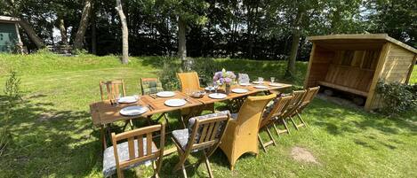 Mesa de madera al aire libre dispuesta para comer, con sillas, rodeada de césped y árboles.