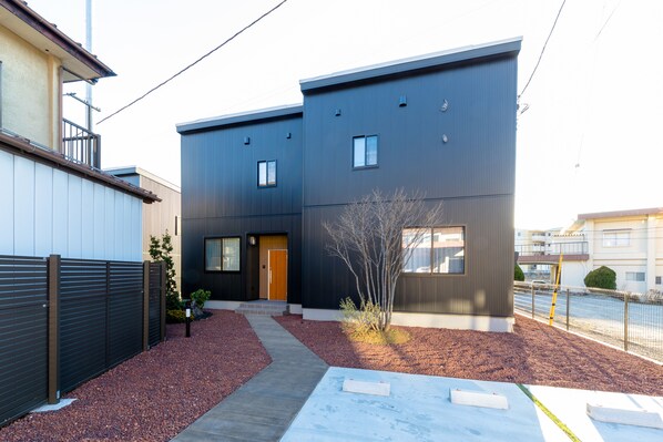 A modern two-story residential building with a dark exterior, a small front yard with a pathway, and a wooden door.