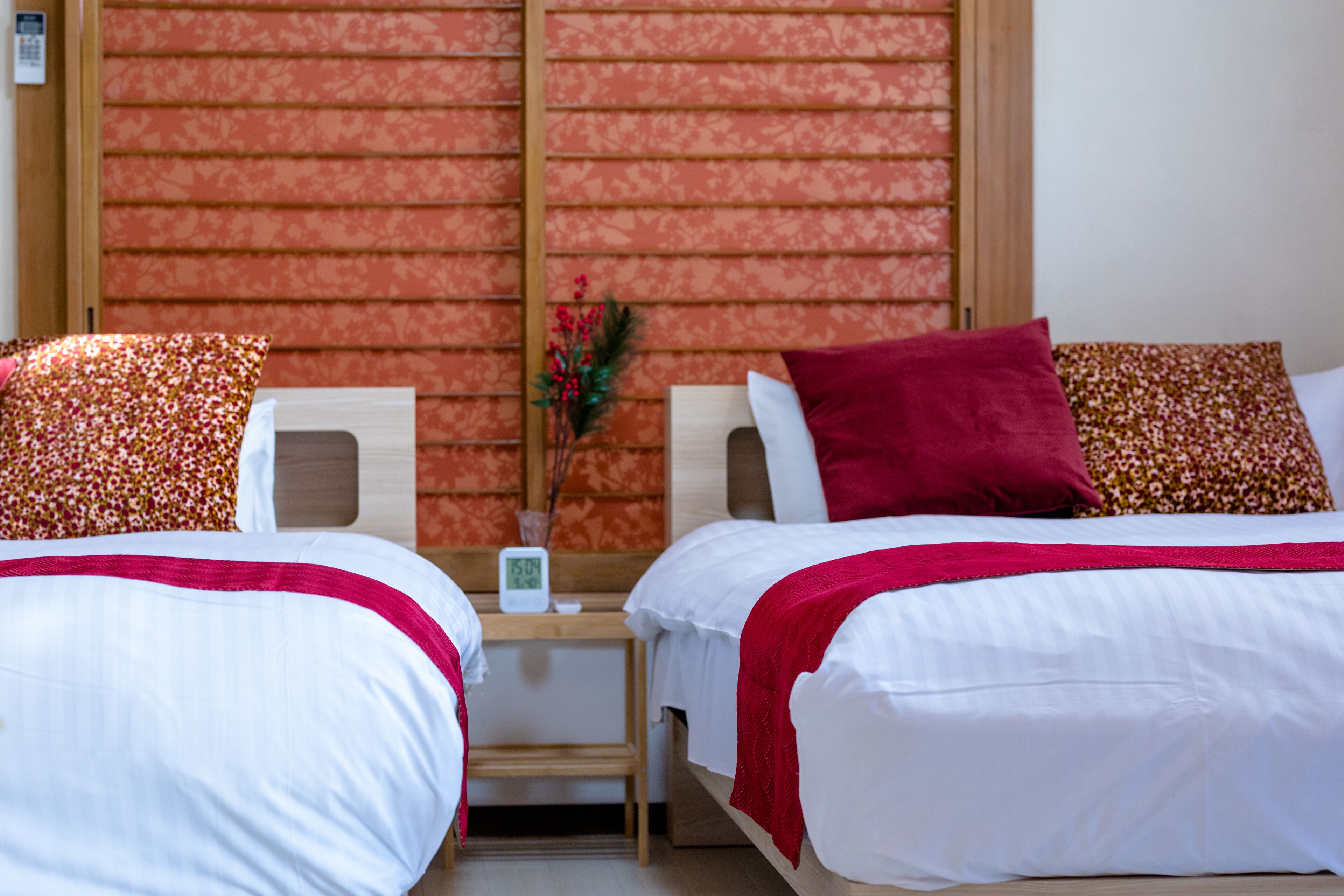 Two single beds with white and red bedding, a small wooden nightstand with a clock, and a decorative plant.