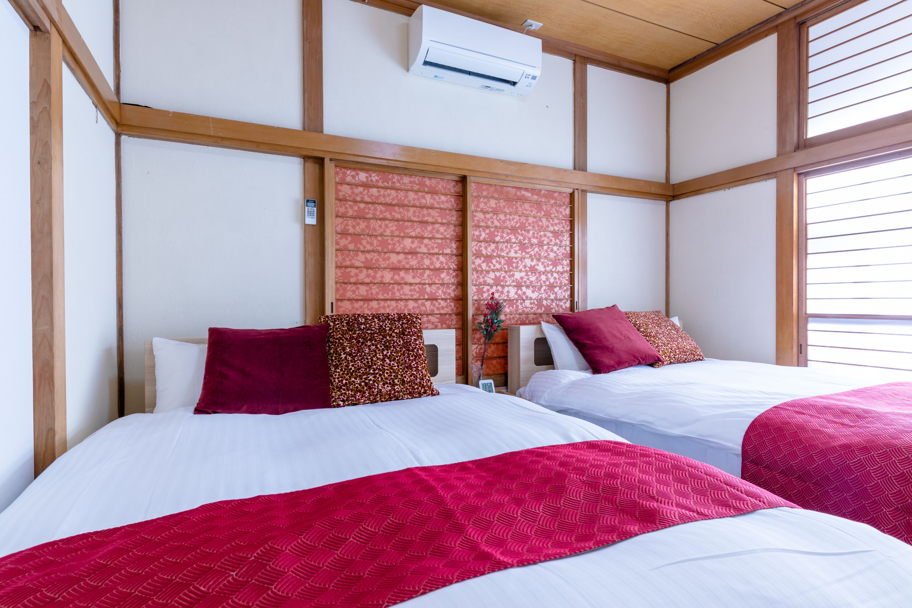 A hotel room with two beds, each with white bedding and red accents, wooden paneling on the walls, and an air conditioning unit above the beds.
