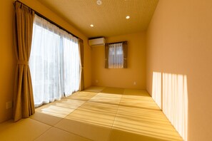 A room with tatami flooring, a sliding door with curtains, a small window with curtains, and an air conditioning unit on the wall.