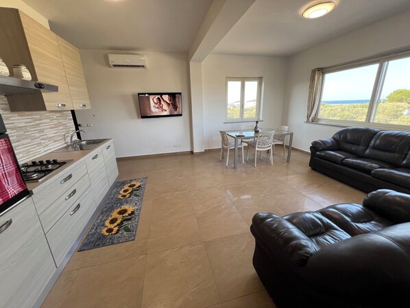 Una sala de estar moderna con un sofá de cuero negro, una mesa de comedor con sillas, una zona de cocina con estufa y un televisor montado en la pared.