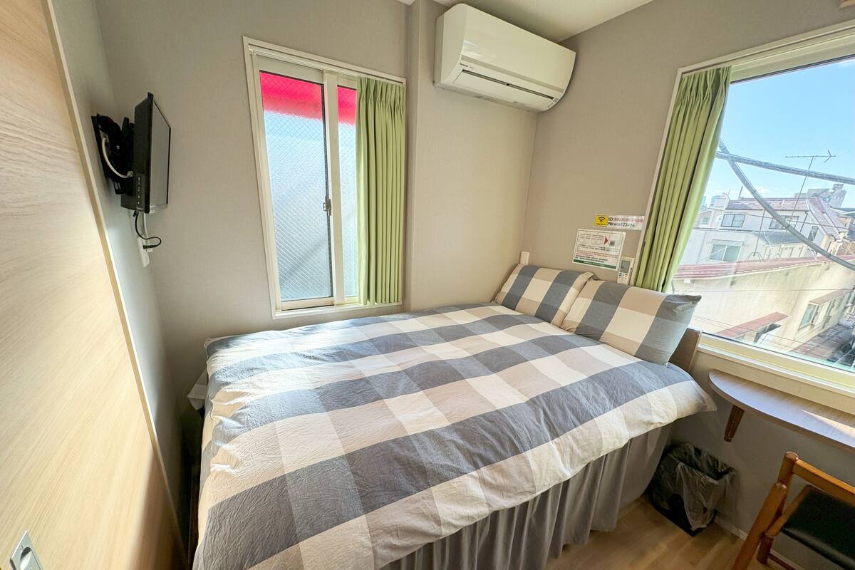 A bedroom with a bed, a window with curtains, an air conditioner, a wall-mounted TV, and a view of buildings outside.