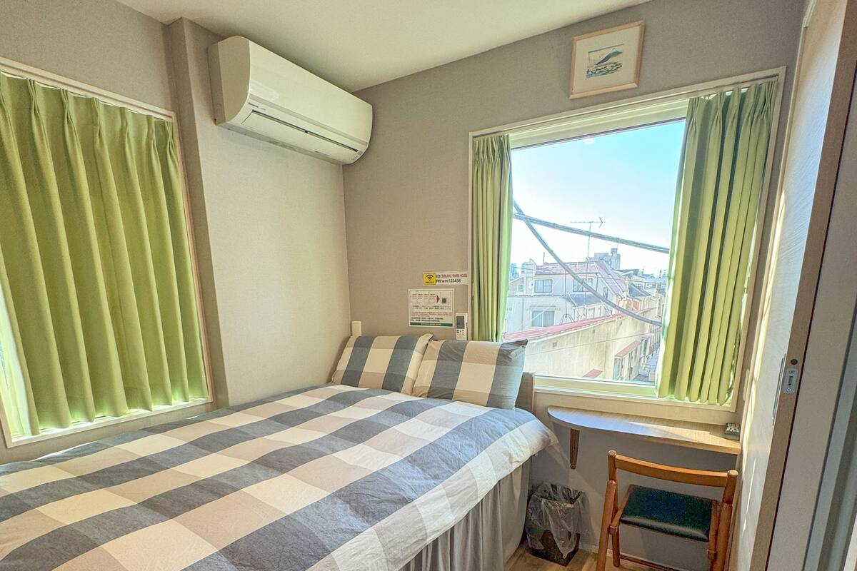 A bedroom with a bed, a desk, and a chair. There is a window with curtains, an air conditioning unit, and a view of buildings outside.