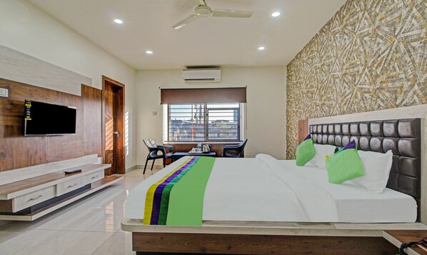A bedroom with a bed, a TV mounted on the wall, a desk with a chair, and a window with a view of buildings. - Treebo Golden Heights (Golmuri)