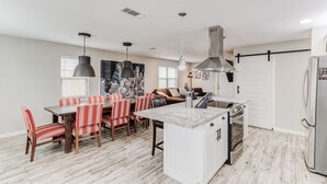 A modern kitchen with a central island, a dining area with striped chairs, and a large window with blinds.