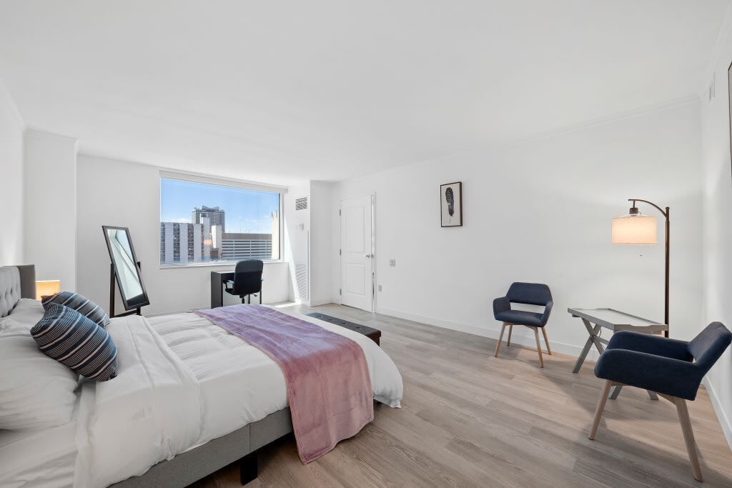 Ein modernes Schlafzimmer mit einem Bett, einem Schreibtisch, zwei Stühlen, einer Stehlampe und einem großen Fenster mit Blick auf die Stadt.