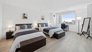 Ein Hotelzimmer mit zwei Betten, einem Schreibtisch, einem Stuhl, einer Stehlampe und einem Fenster mit Blick auf die Stadt.