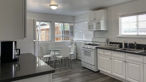 Una cocina moderna con armarios blancos, estufa a gas, microondas y cafetera. Hay una mesa de comedor con sillas y una ventana con persianas.