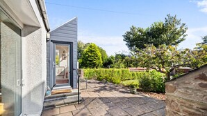 A house with a grey exterior, a door leading to a small porch, and a garden with hedges and trees. - The Albatross - 3 bed Carnoustie Classic (Angus)