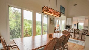 A dining area with a wooden table and wicker chairs, large windows with a view of greenery, and a kitchen in the background. - 2 Minute Walk to Beach; Access to Community Pool and Pickle Ball, Basketball & Tennis Courts (Isle of Palms)