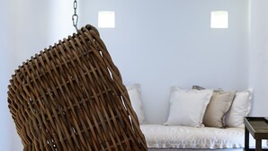Une chaise suspendue faite de branches entrelacées, un canapé blanc avec des coussins et un petit tabouret en bois, dans une pièce aux murs blancs et au sol carrelé.