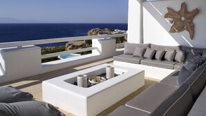 Un espace extérieur moderne, aménagé avec un canapé et des coussins blancs, un foyer et une vue sur la mer.