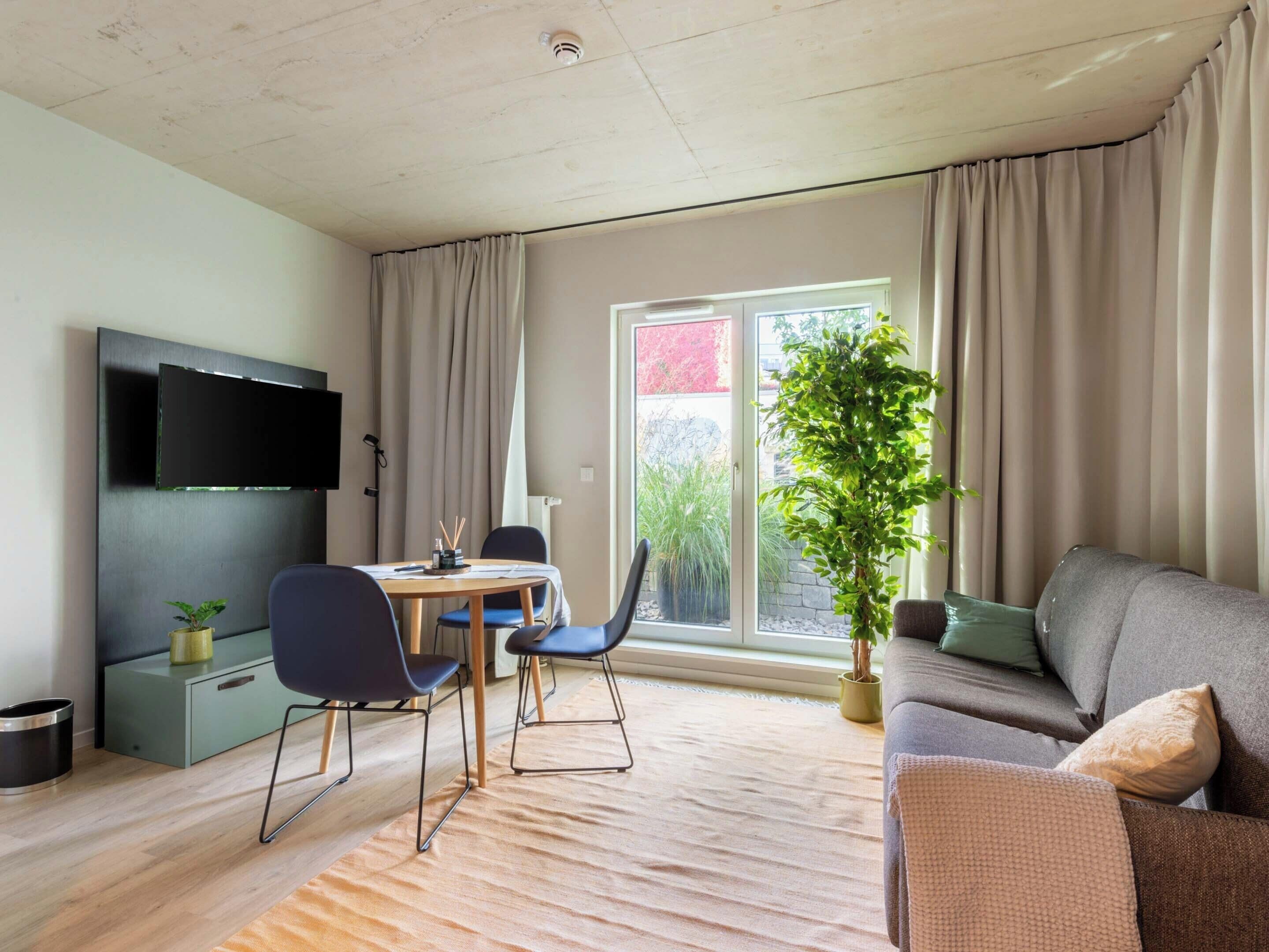A modern living room with a flat-screen TV, a dining table with chairs, a sofa, and a large window with curtains.
